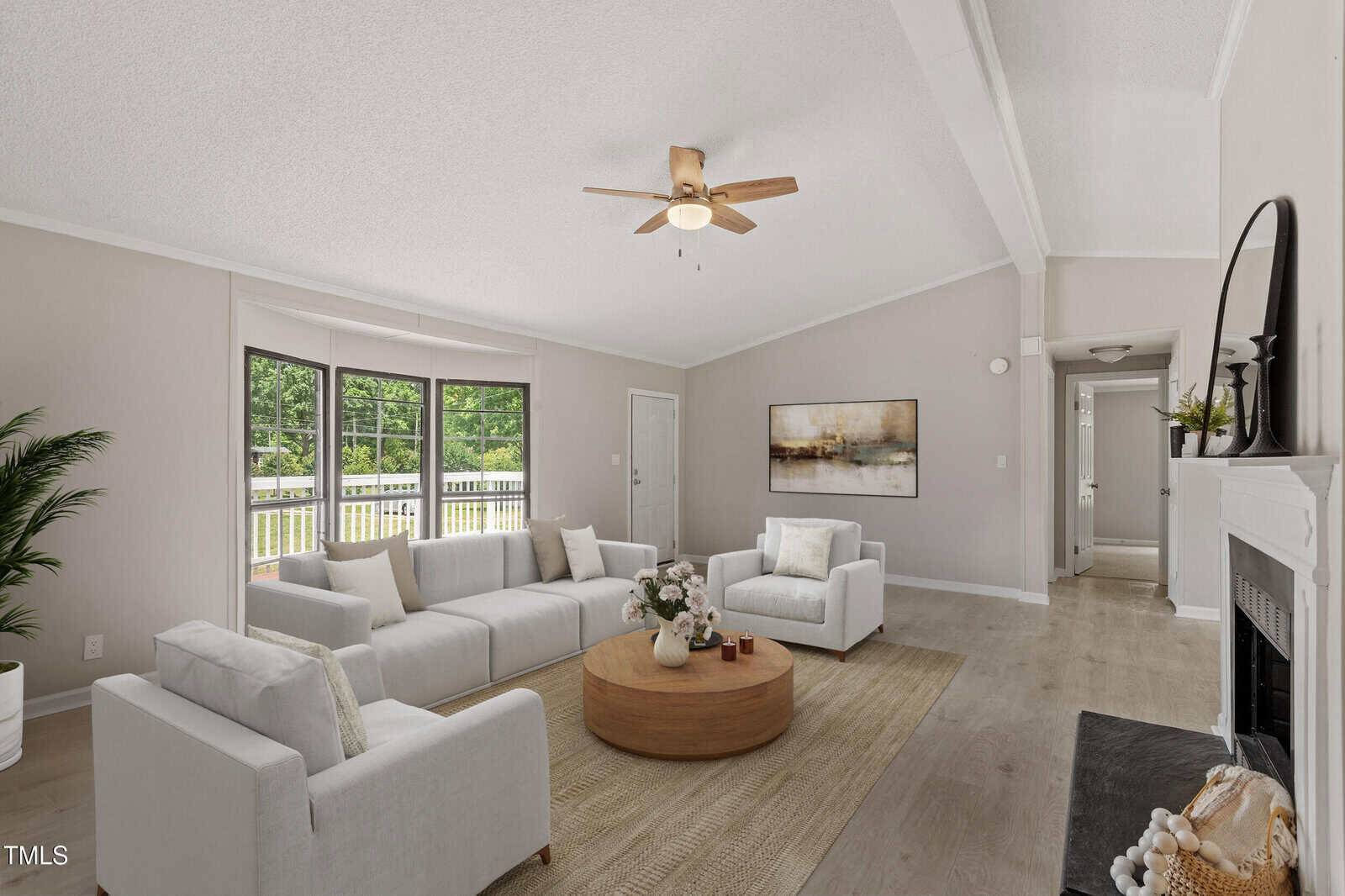 A bright living room with light walls and floors, white sofas, a round wooden coffee table, a ceiling fan, a fireplace, and large windows showing greenery outside. Neutral decor creates a cozy, modern atmosphere.