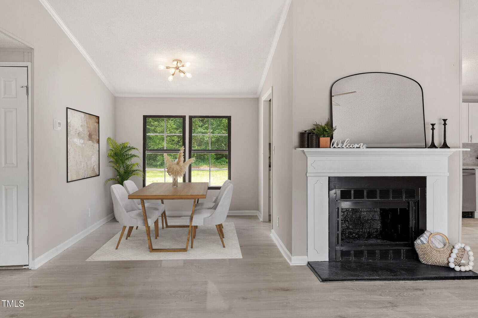 A dining area with a wooden table, four upholstered chairs, a plant, and a decorative fireplace with a mirror and basket. Two large windows provide natural light. The room features light walls and hardwood floors.