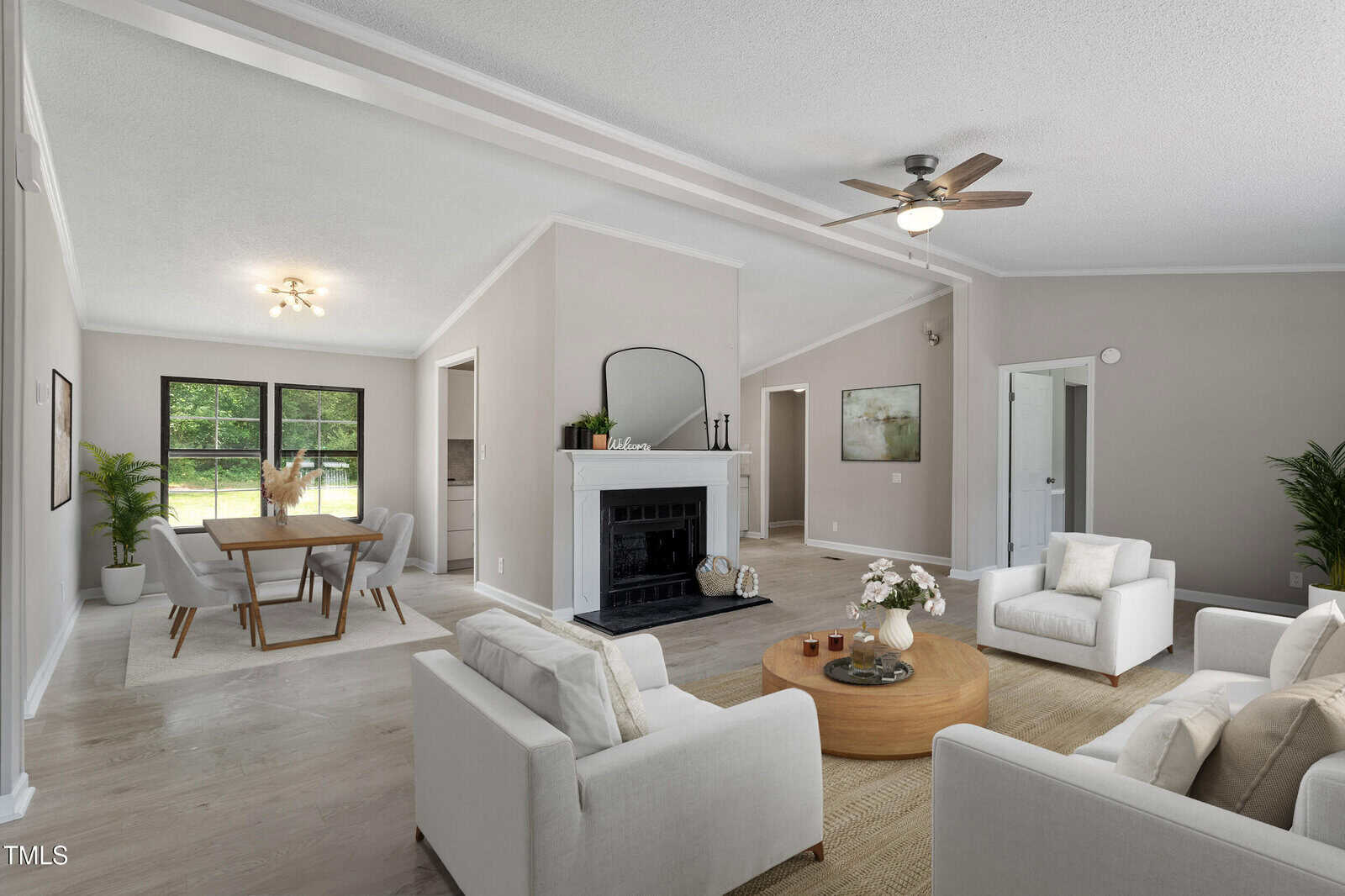 A modern living and dining area with light-colored walls and floors, white sofas, a round wooden coffee table, a white fireplace with a mirror above, and large windows letting in natural light.