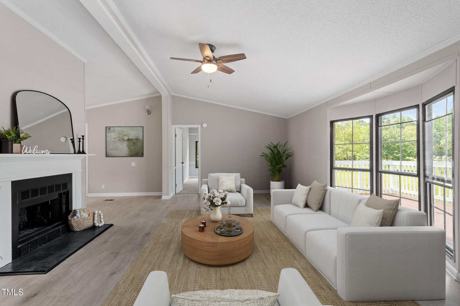 A modern living room with light gray walls, two white sofas, a round wooden coffee table, a fireplace with a mirror above, large windows, a ceiling fan, and a potted plant in the corner.