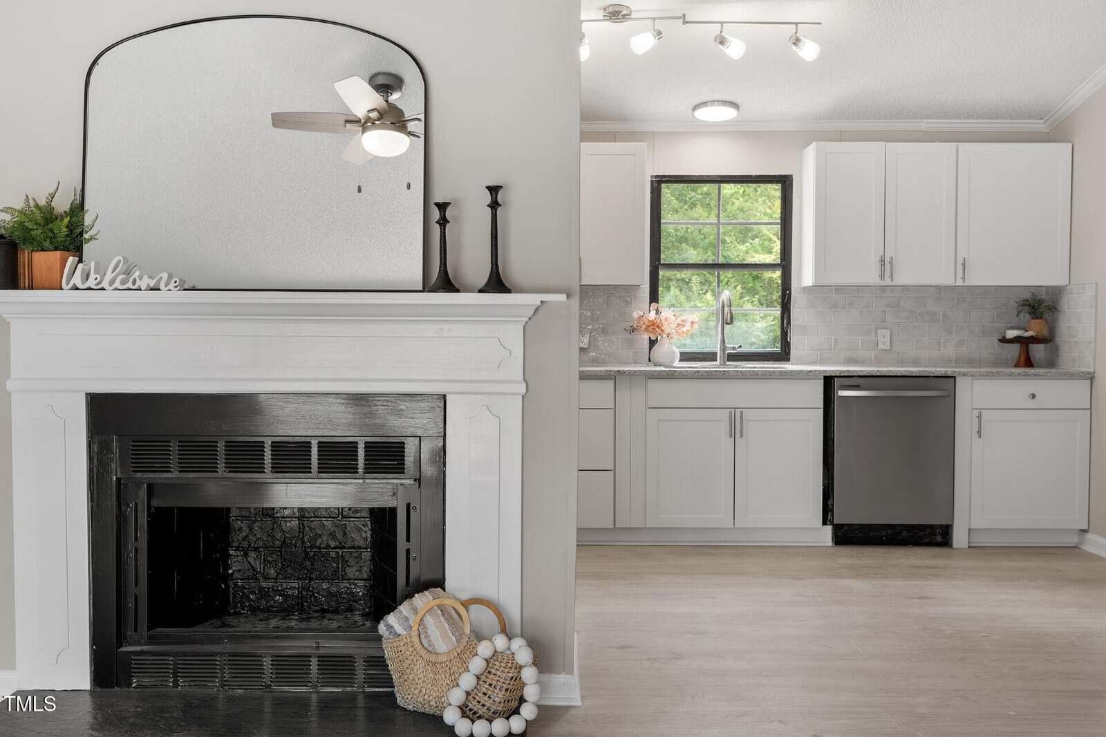 A modern kitchen with white cabinets and stainless steel appliances is visible next to a white fireplace with a black interior, a large mirror, candlesticks, a "Welcome" sign, and decorative items on the floor.