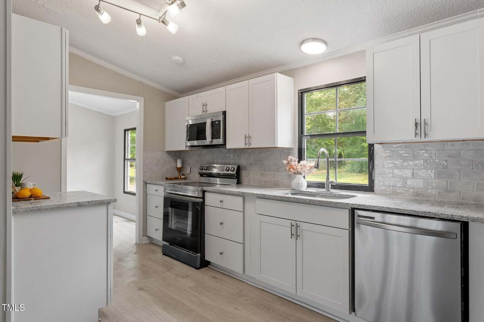 Modern kitchen with white cabinets, stainless steel appliances, gray subway tile backsplash, light granite countertops, and large windows letting in natural light. A bowl of fruit and vase with flowers sit on the counters.