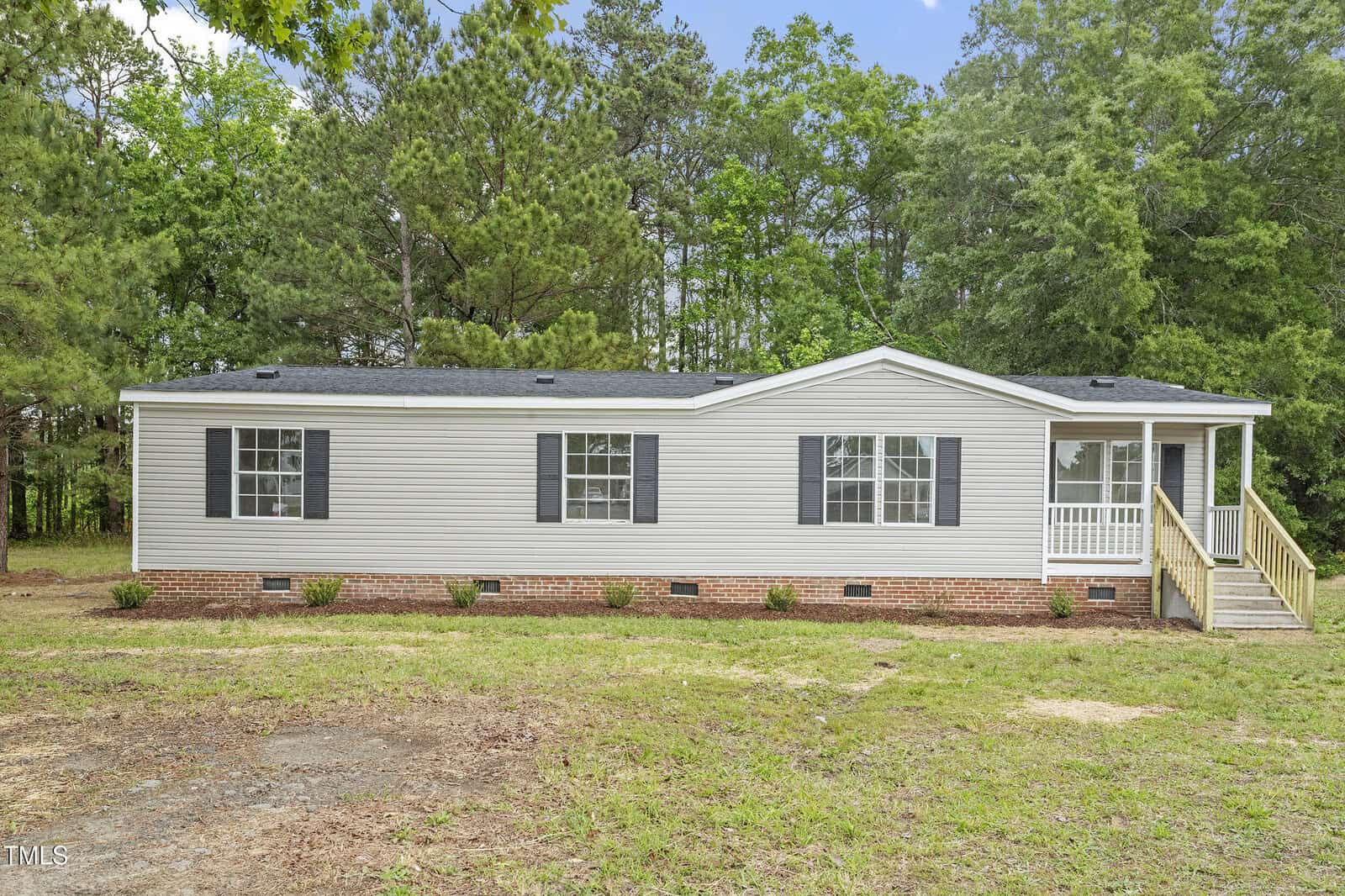 Located at 2089 Lisa Lane in Creedmoor, this single-story manufactured home features beige siding, black shutters, and a small covered front porch. The house sits on a brick foundation with grass, sparse landscaping, and trees in the background.