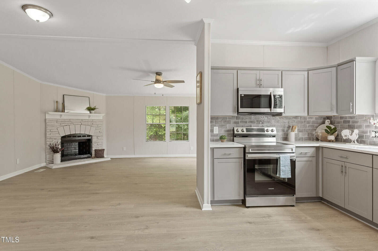 A modern kitchen with gray cabinets, stainless steel appliances, and a gray tile backsplash is shown next to an open living area with light wood floors, a white brick fireplace, and large windows.