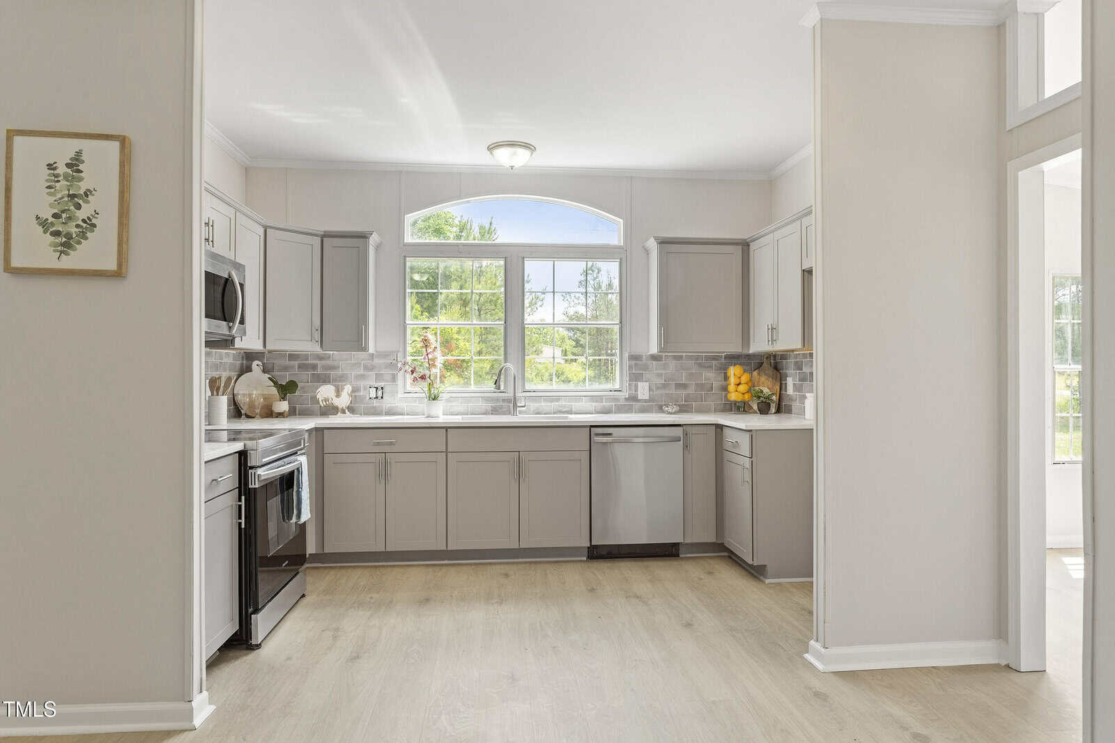 A modern kitchen with light gray cabinets, stainless steel appliances, white countertops, a large window above the sink, light wood flooring, and a framed botanical print on the wall.