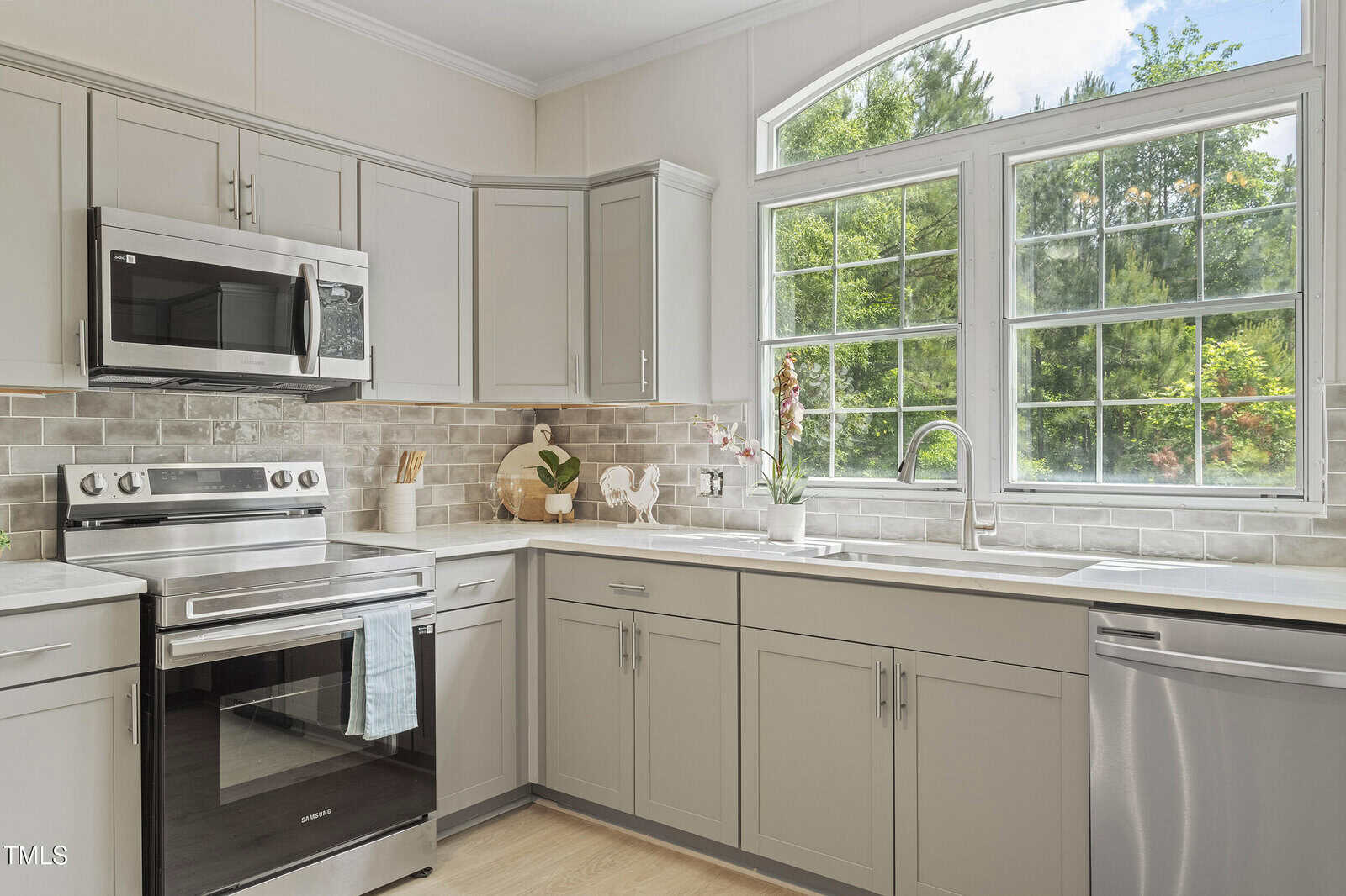 Modern kitchen with light gray cabinets, stainless steel appliances, and a large window above the sink. The countertop is white, and the backsplash features light-colored subway tiles. Natural light fills the space.