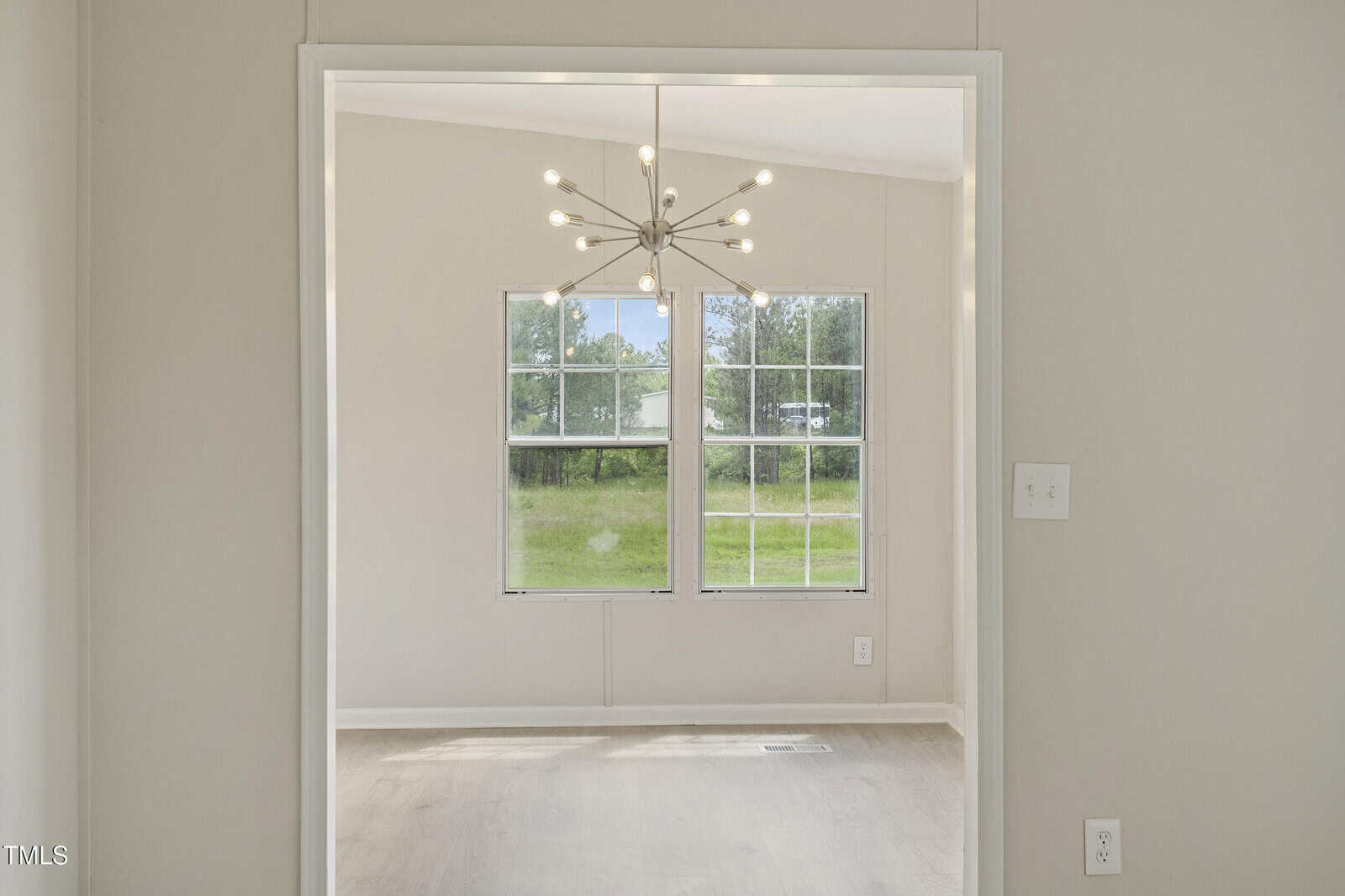 A white-framed interior doorway opens to a room with two large windows, a modern sputnik chandelier, light wood flooring, and a green outdoor view. Light switches are visible on the wall to the right.
