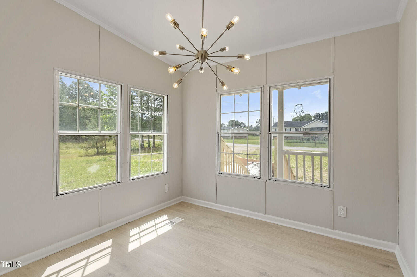 A bright room with three large windows, light wood flooring, off-white walls, and a modern chandelier hanging from the ceiling. Trees and a house are visible outside through the windows.