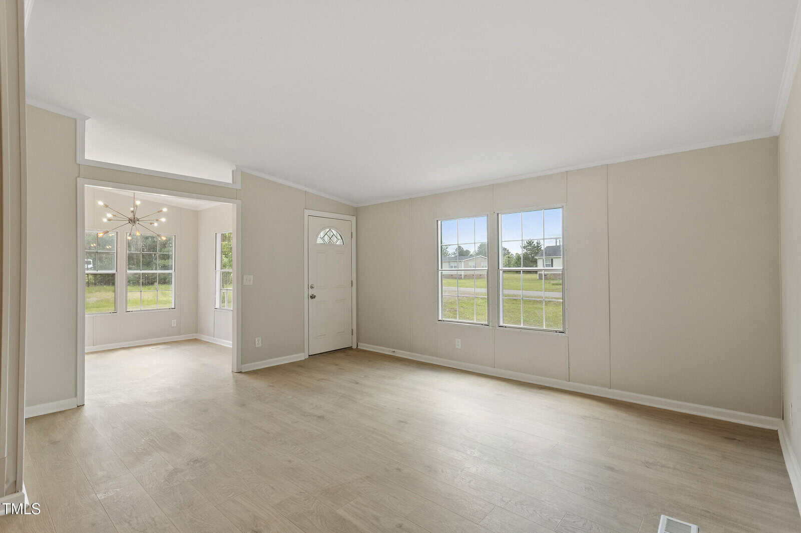 A bright, empty living room with light-colored walls, wood flooring, two large windows, and a white front door. An adjacent room with more windows and a modern chandelier is visible in the background.