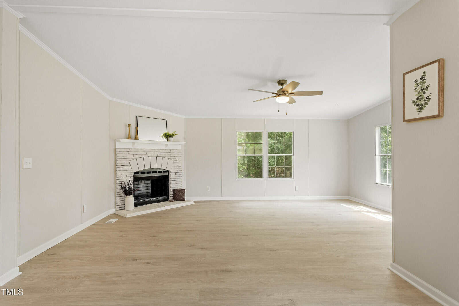 A spacious, empty living room with light wood flooring, white walls, a white brick fireplace, ceiling fan with light, large windows letting in natural light, and a framed plant print on the wall.