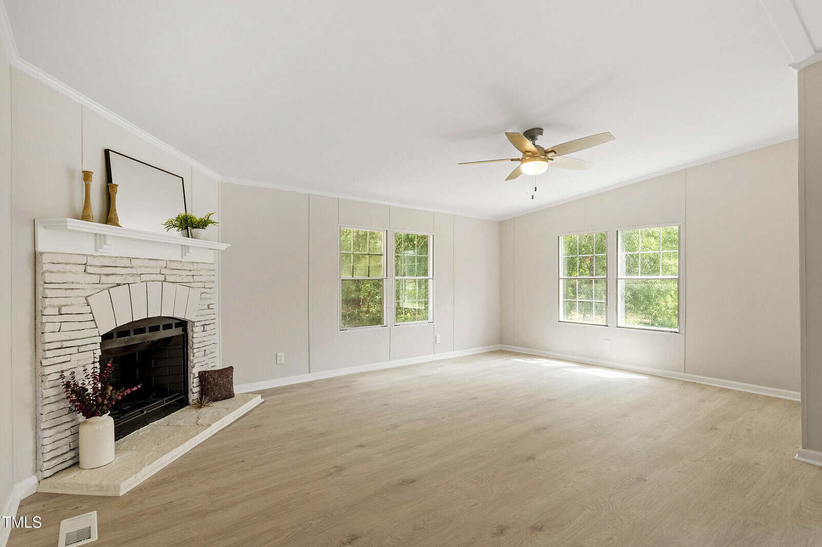A bright, empty living room with light wood floors, white walls, three large windows, a ceiling fan, and a white brick fireplace with a mantel decorated with a mirror and vases.