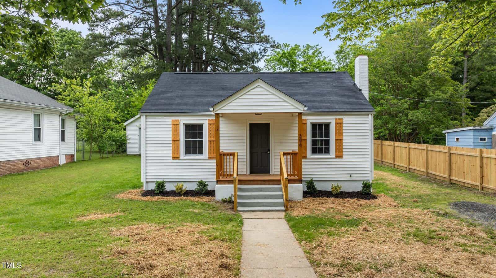 Single-story white house with a small covered front porch, wooden shutters, and a gray roof. The house is surrounded by a grassy yard, with a sidewalk leading to the front steps and trees in the background.