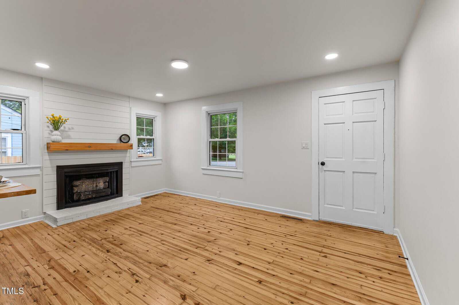 A bright living room with wood floors, white walls, recessed lighting, a white-painted brick fireplace with a wood mantel, two windows, and a white door. The room has minimal decor and a clean appearance.