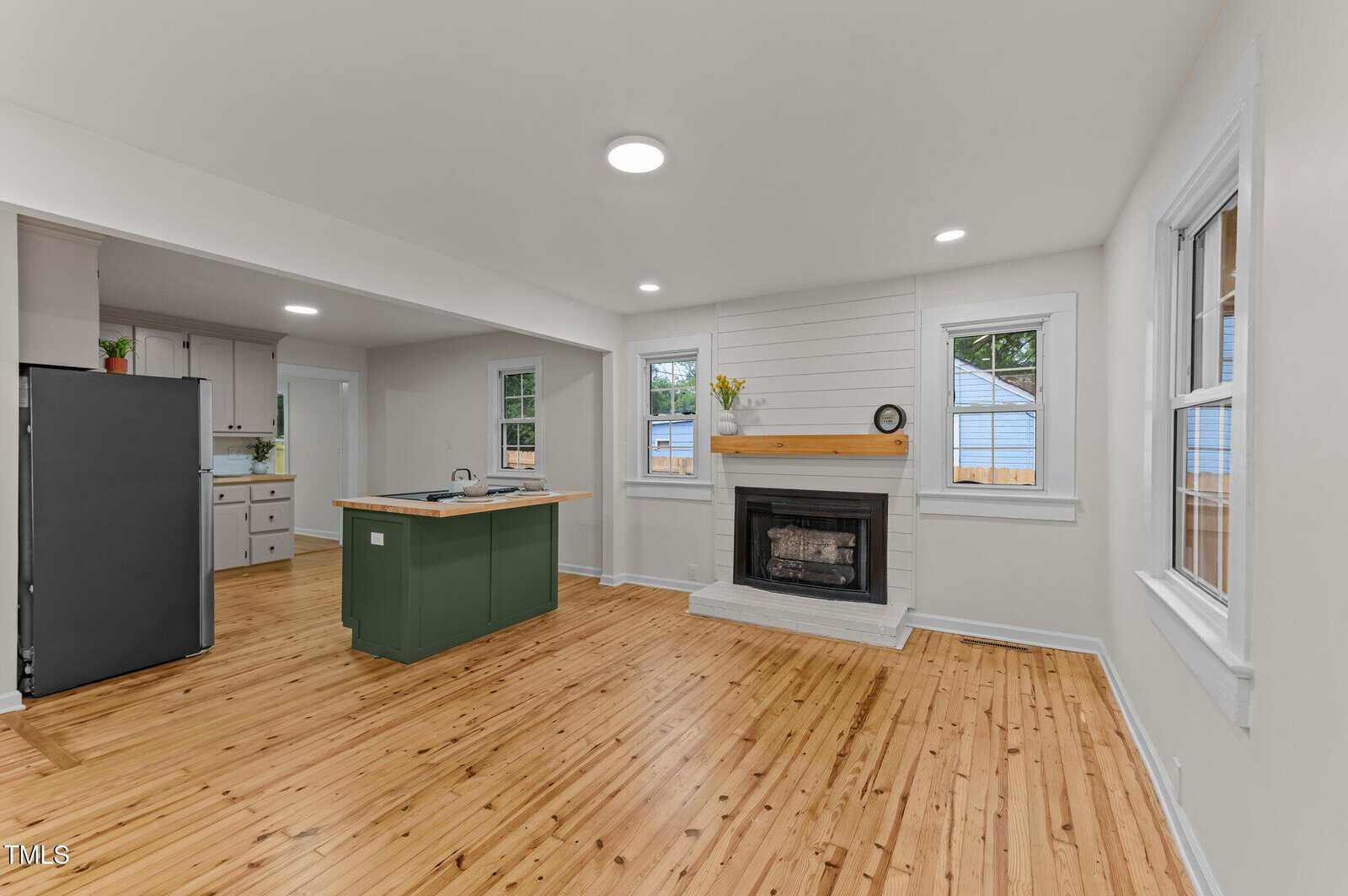 A bright, open-concept kitchen and living area with light wood flooring, a green kitchen island, stainless steel refrigerator, white walls, and a fireplace with a wooden mantel and shiplap accent. Multiple windows provide natural light.