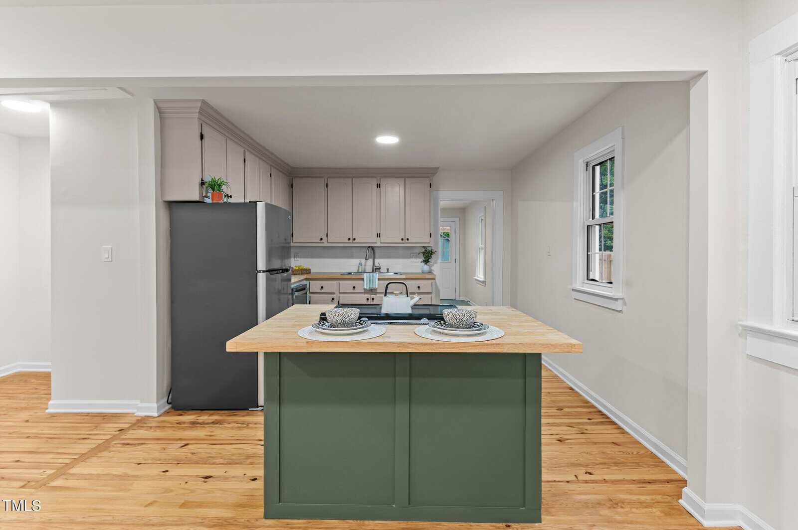 A kitchen with light-colored walls, wood floors, a green island with place settings for two, stainless steel refrigerator, white cabinets, and a window on the right. The space is brightly lit with recessed ceiling lights.