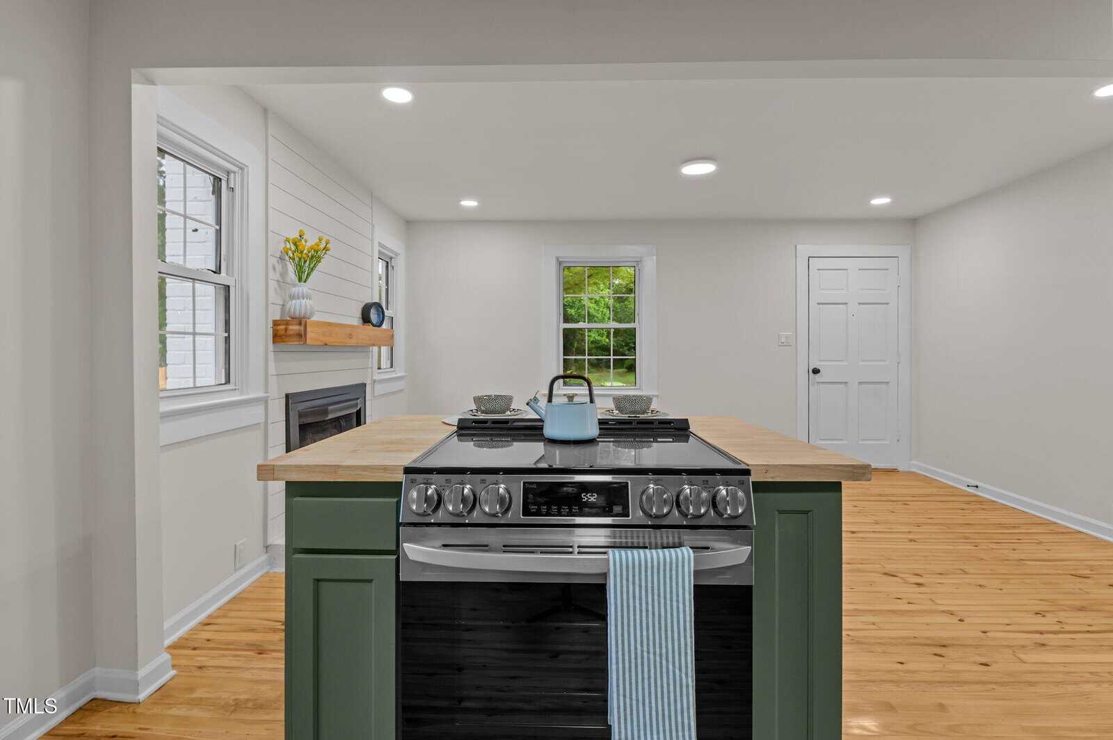 A modern kitchen with a central island featuring a green base, wooden countertop, gas stove, and blue kettle. The room has light-colored walls, hardwood floors, and large windows letting in natural light.