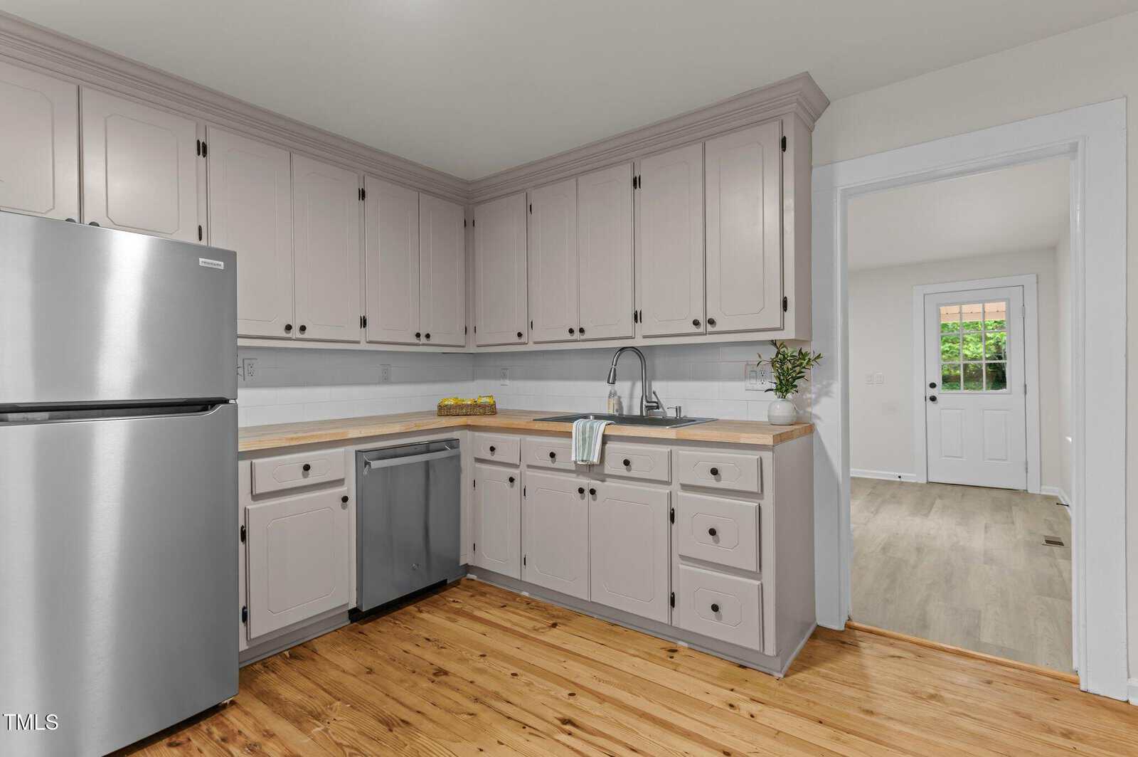 A kitchen with light beige cabinets, stainless steel fridge and dishwasher, wood countertops, white tile backsplash, and a wood floor. A door leads to another room with light-colored flooring. A towel hangs by the sink.