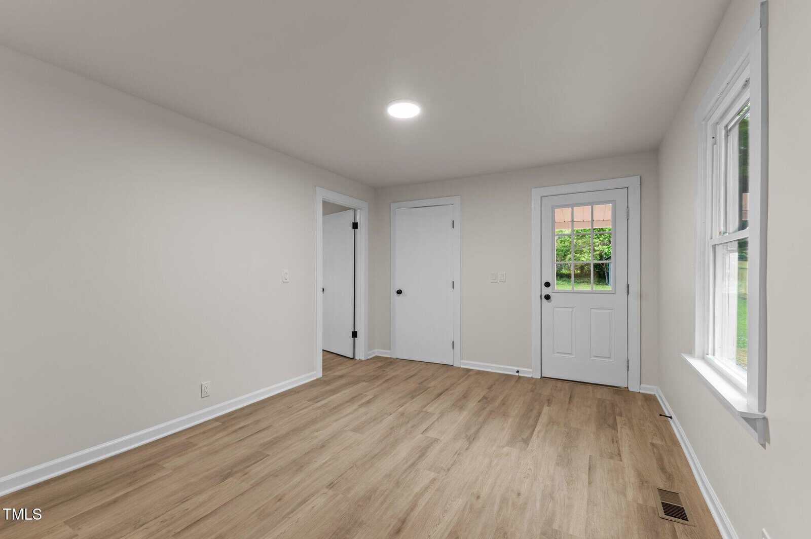 Empty room with light-colored walls, wood-style flooring, a white door with a window, two closed white doors, and a window showing greenery outside; ceiling light fixture is on.