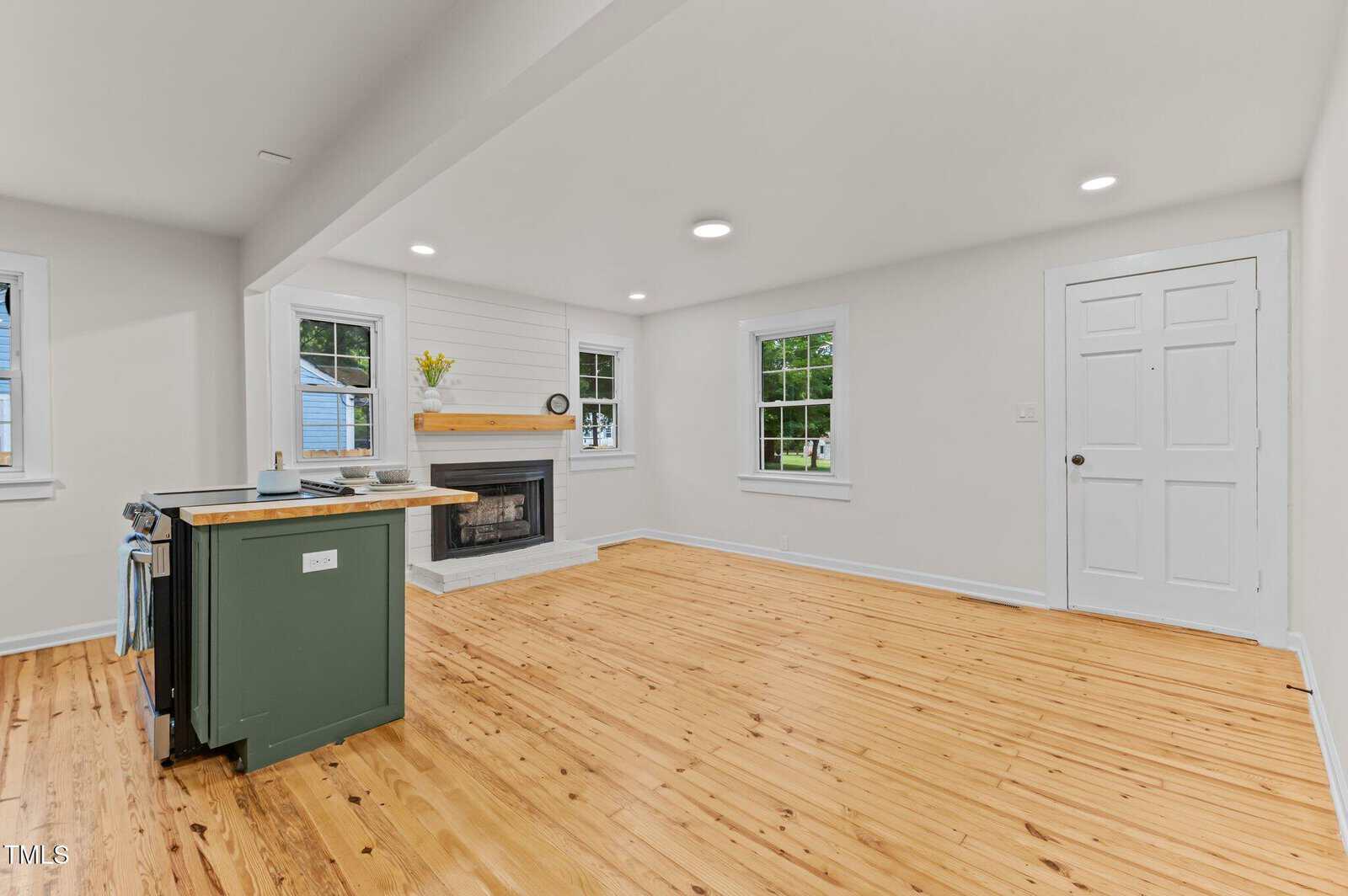 A bright, unfurnished living space with light wood flooring, white walls, recessed lighting, a green kitchen island, a fireplace, and several windows. A white door is on the right side of the room.