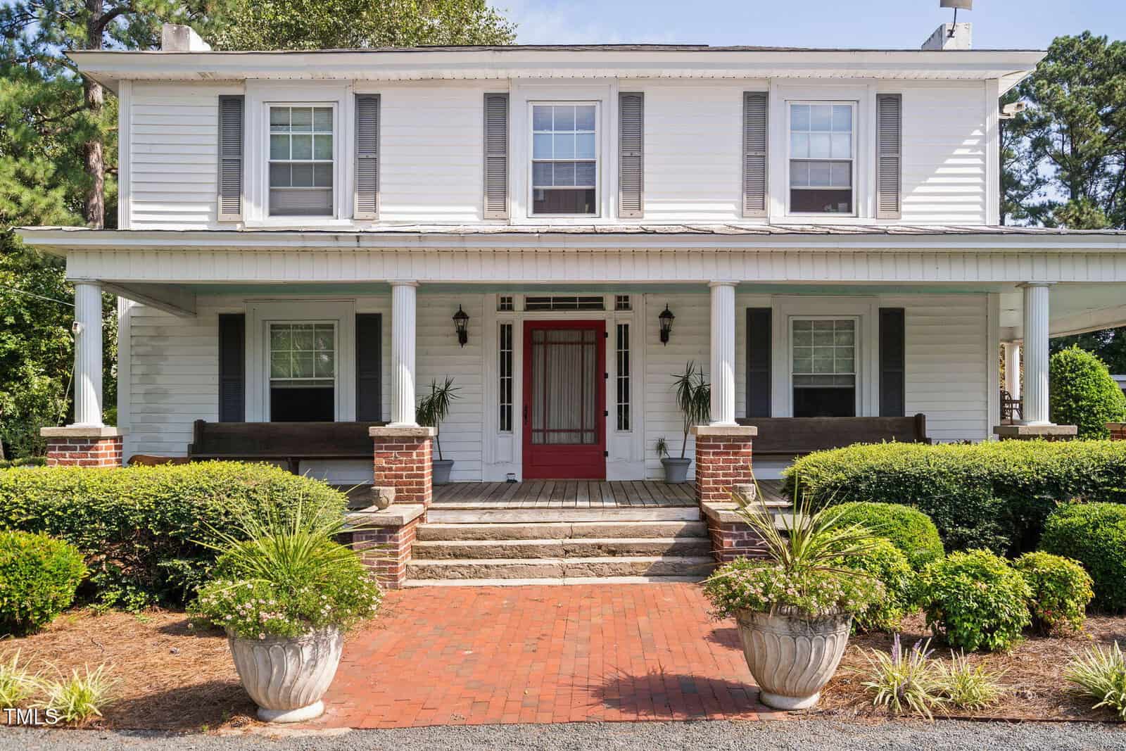 A two-story white house with gray shutters, a red front door, a covered porch supported by white columns, brick steps, planters with greenery, and manicured bushes in front.