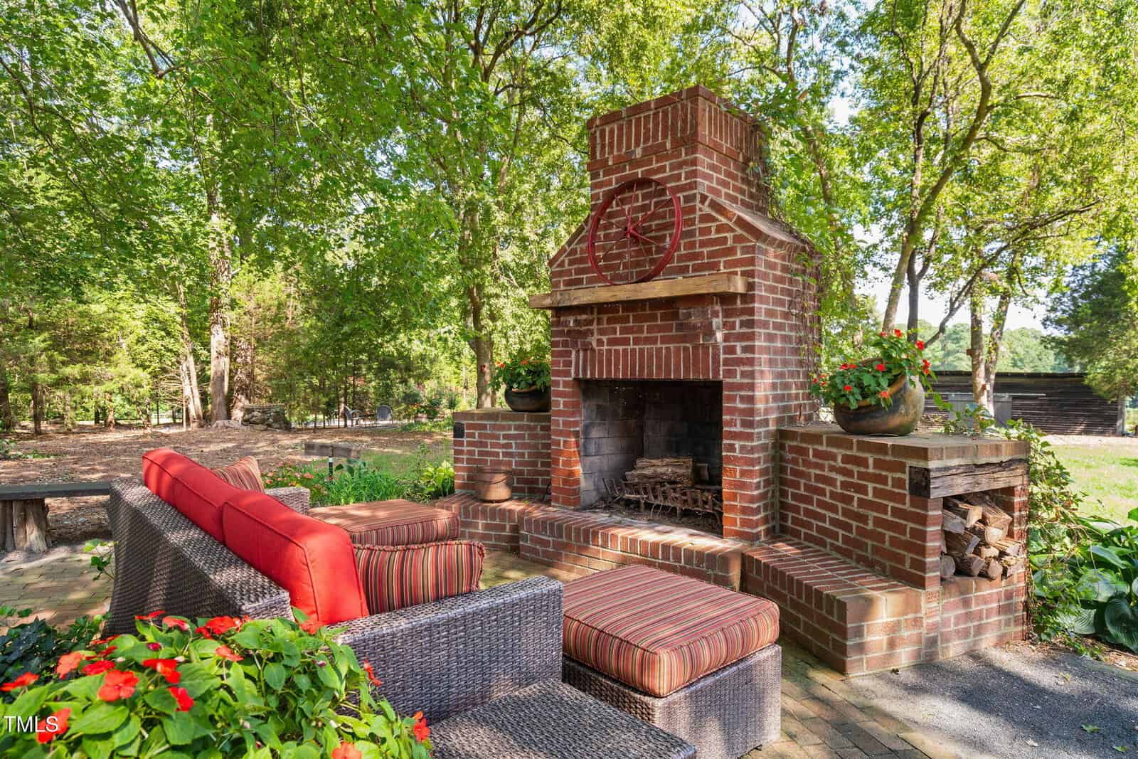 An outdoor patio with a large brick fireplace, wicker furniture with red cushions, potted flowers, and firewood stored on the side, surrounded by trees and greenery on a sunny day.