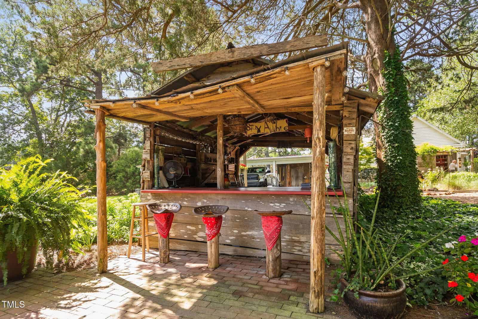 A rustic outdoor bar with a wooden roof and three stools, each with a red bandana. The area is surrounded by greenery, potted plants, and brick flooring, with a fan and decor inside the bar.
