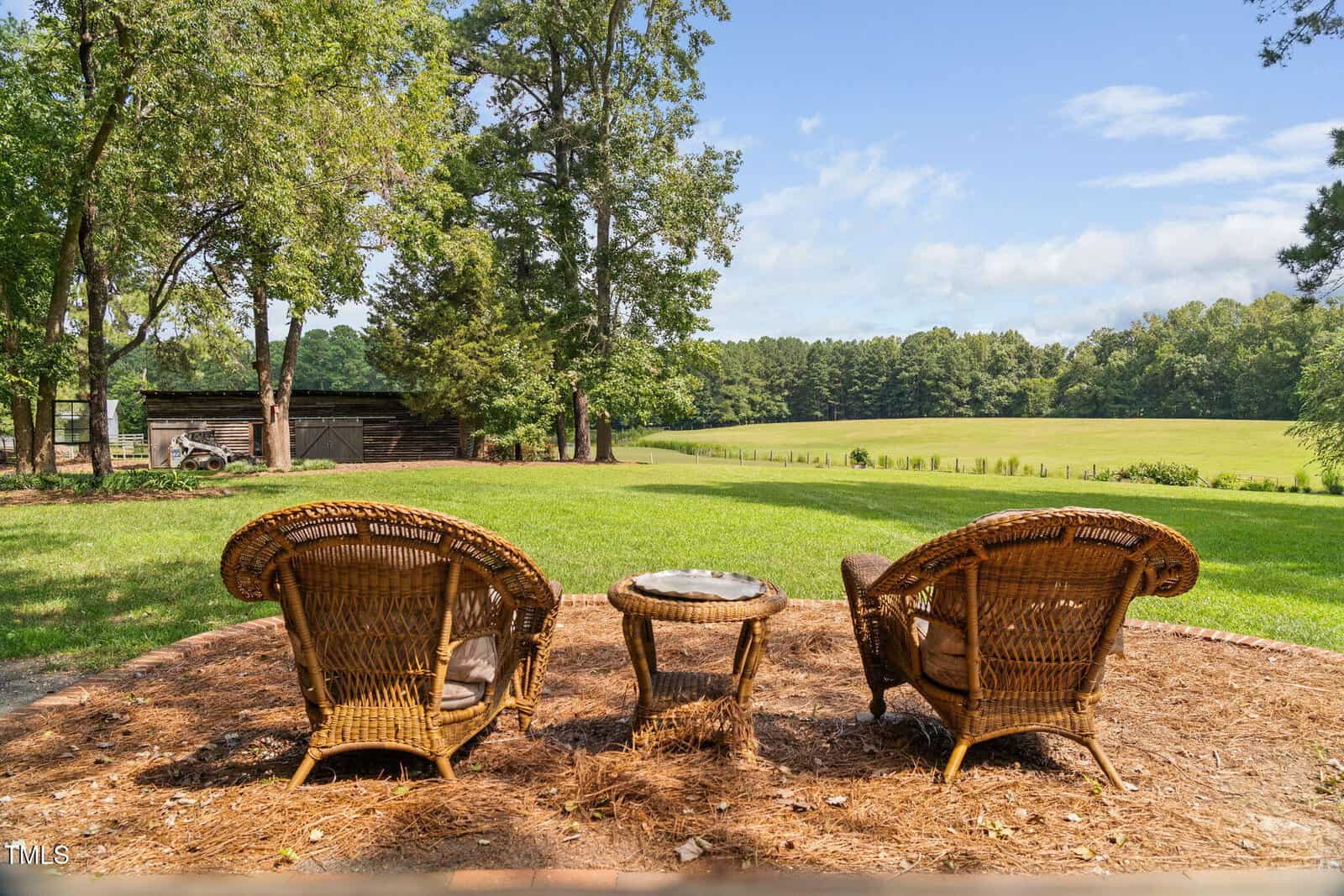 Three wicker chairs and a small table are arranged in a semicircle on a patch of pine straw, overlooking a grassy lawn, trees, and an open field under a partly cloudy sky. A wooden shed stands in the background to the left.