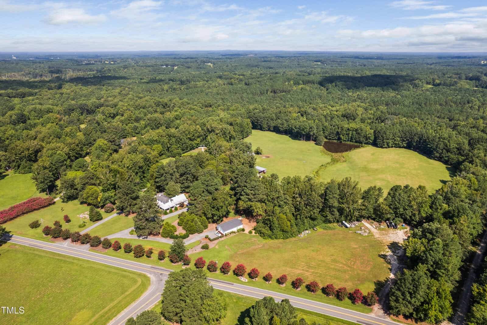 Aerial view of a rural property with several houses, open fields, fenced areas, a small pond, a tree-lined road, and dense forest surrounding the area under a partly cloudy sky.