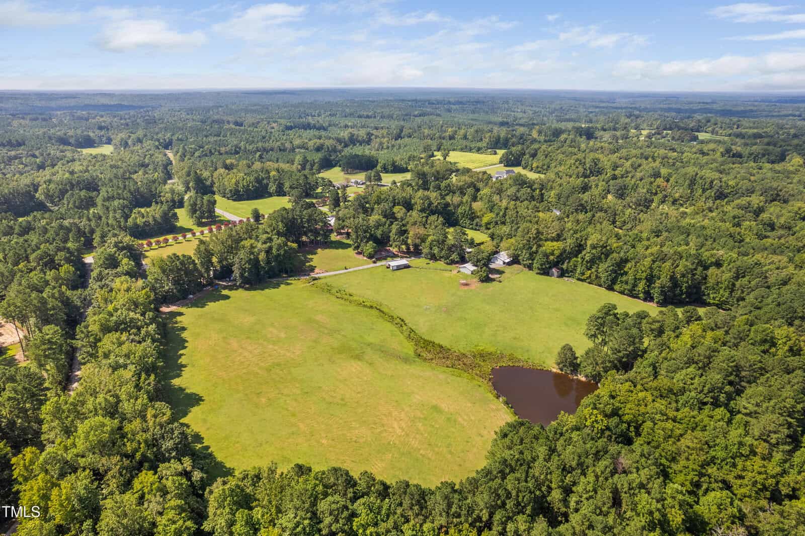 Aerial view of a large, open grassy field bordered by trees, with a pond in the lower right and several buildings and roads scattered throughout the landscape under a blue sky with clouds.