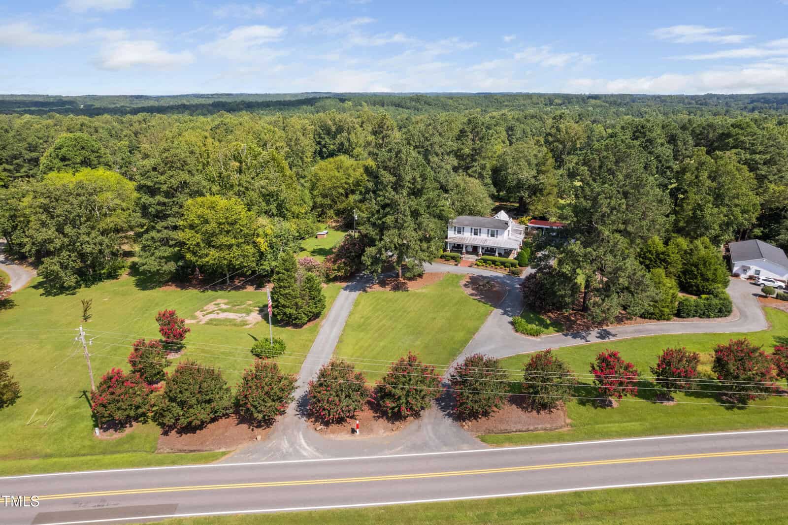 A large white house with a red roof sits among trees on a spacious, well-manicured property with long driveways, neatly trimmed lawns, and red-flowering shrubs along a two-lane road. Dense forest surrounds the area.