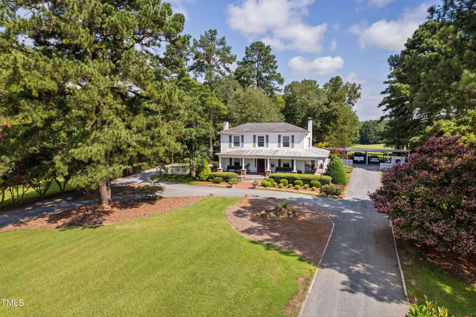 A two-story white house with a covered front porch sits near the center of a large, well-kept lawn surrounded by tall trees. A circular driveway leads to the front entrance, and several cars are parked in the background.