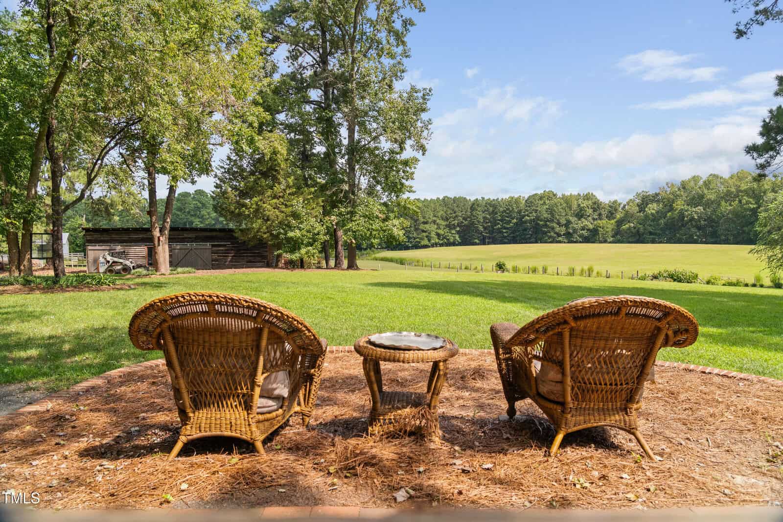 Two wicker chairs and a small table sit on a mulched area overlooking a green lawn and open field with trees and a barn in the background under a partly cloudy sky.