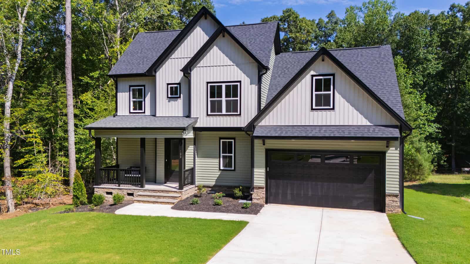 Two-story modern suburban house with gray siding, black trim, covered front porch, and attached two-car garage. The home features a well-kept lawn, concrete driveway, and is surrounded by trees and greenery.
