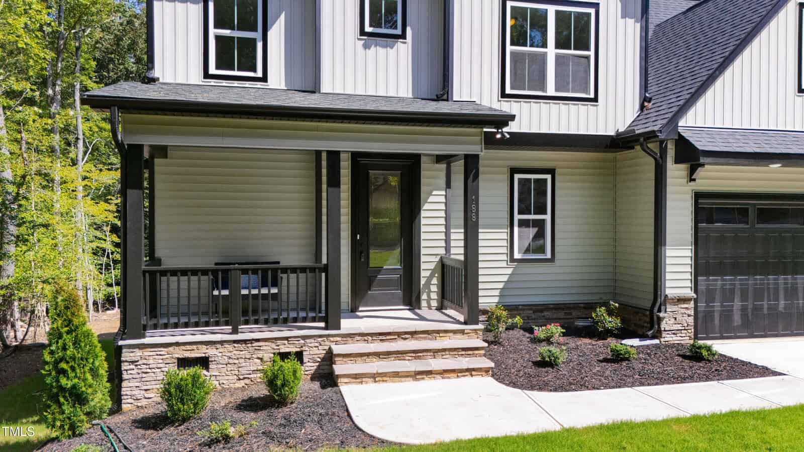 A modern house exterior with light-colored siding, black trim, a small covered front porch with railings, stone accents, steps leading to the door, landscaped mulch beds, and a green lawn.