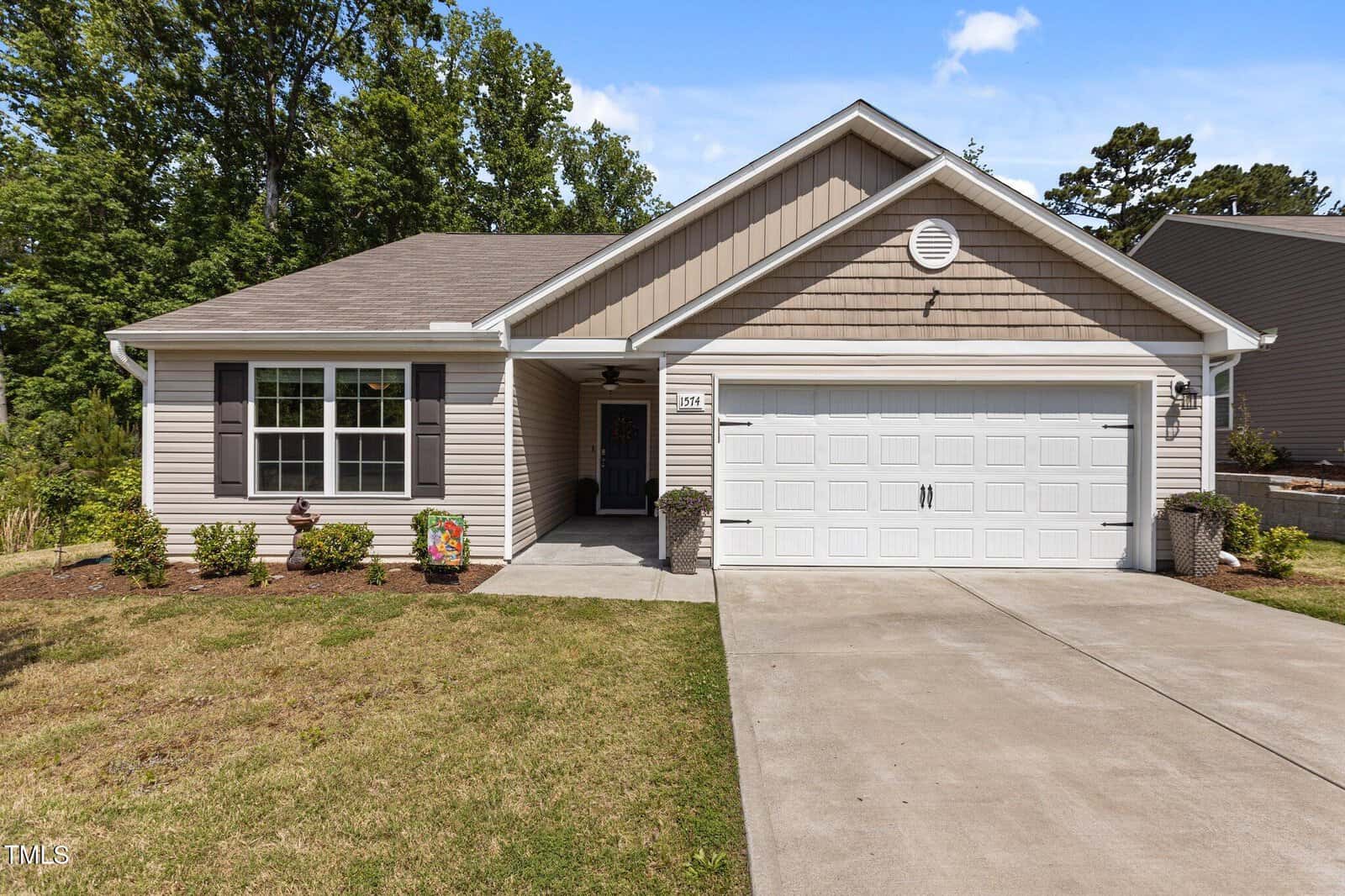 Charming single-story suburban house at 1574 Dorsett Lane, Creedmoor, featuring beige siding, brown accent shingles, white trim, and a two-car garage. A concrete driveway leads to the entrance with simple landscaping and trees in the background.