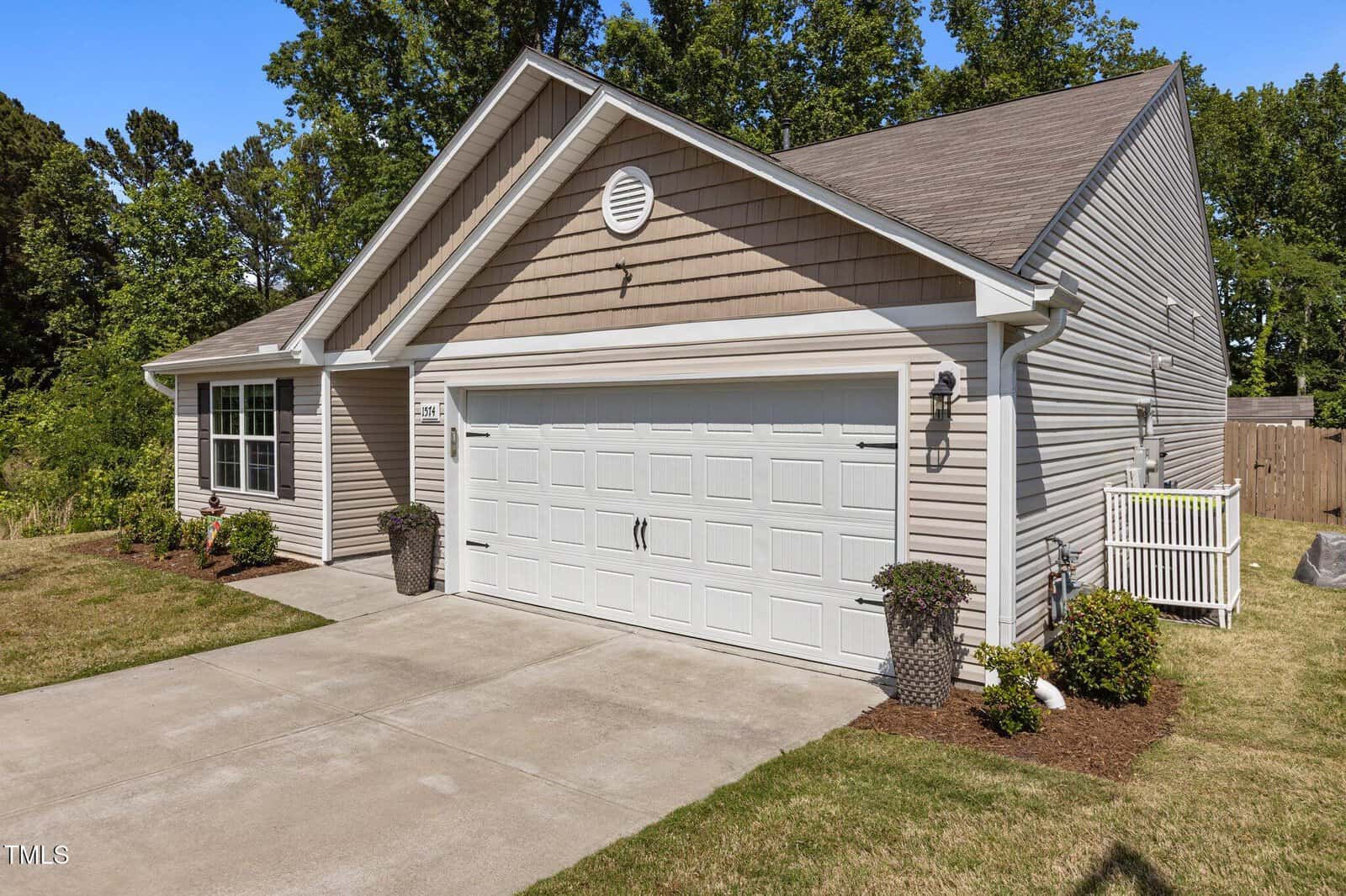 A single-story suburban house with beige siding, a two-car white garage door, a concrete driveway, small landscaped bushes, and a fenced backyard, set against a backdrop of green trees.