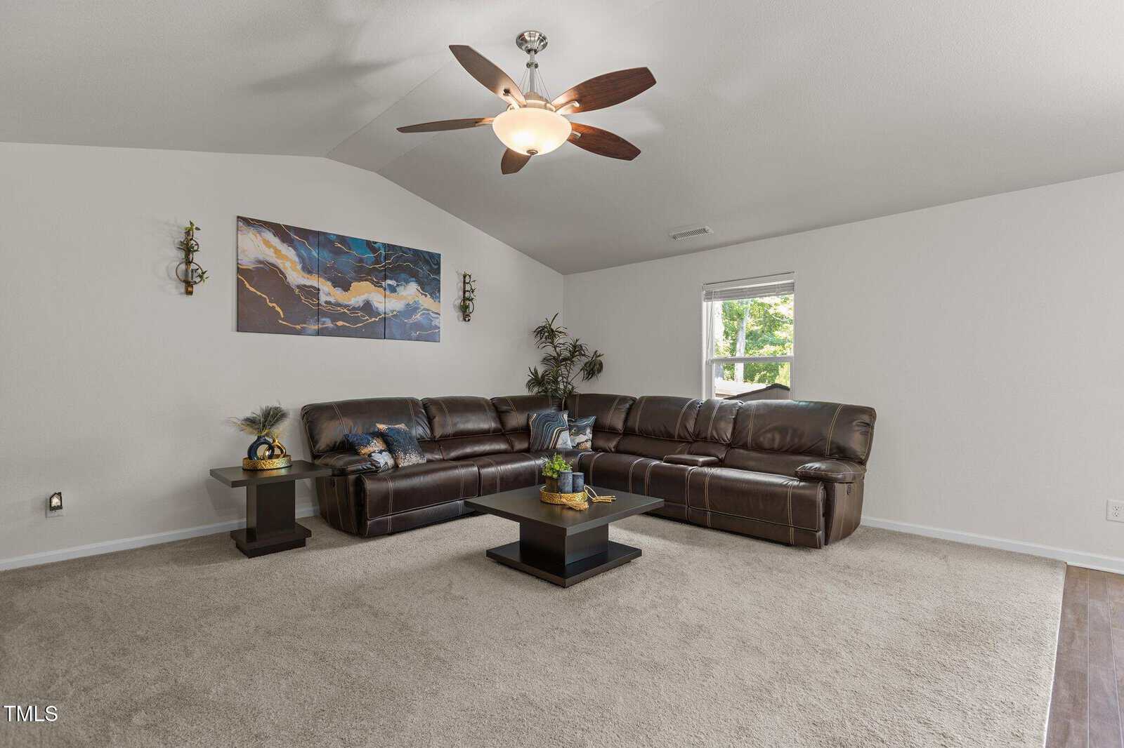 A living room with a brown leather sectional sofa, a modern ceiling fan, a window with sunlight, abstract wall art, two side tables with plants and decor, and a central coffee table on beige carpet.