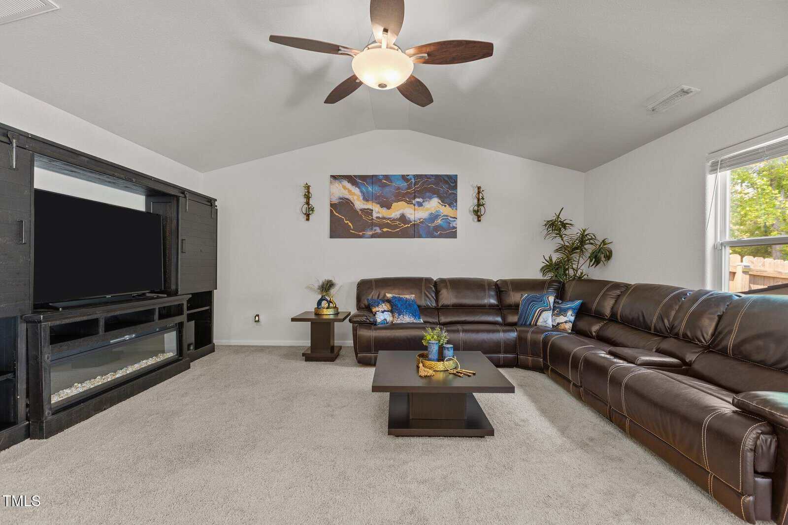 A spacious living room with a large brown leather sectional sofa, a square coffee table, a flat-screen TV on a black entertainment unit, wall art, potted plants, and a ceiling fan with a light. Natural light comes from a window.