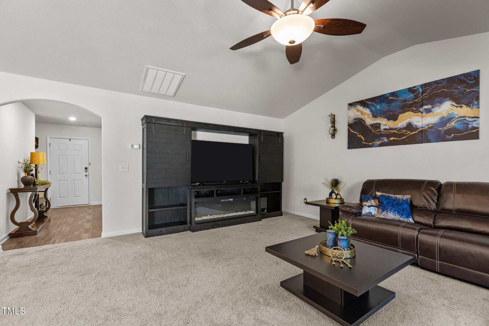 A living room with beige carpet, a dark entertainment center with a TV, a brown leather sofa, a modern coffee table with plants, a blue and gold abstract painting, and a ceiling fan with lights.