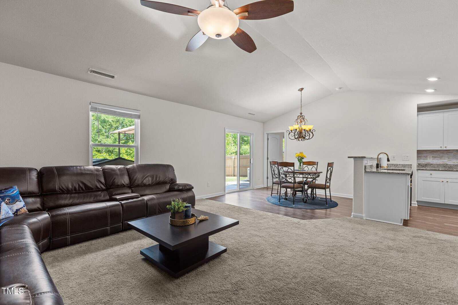 Spacious open-concept living and dining area with brown leather sectional sofa, square coffee table, carpeted floor, chandelier over dining table, and view of kitchen with bar counter; large windows and door show greenery outside.