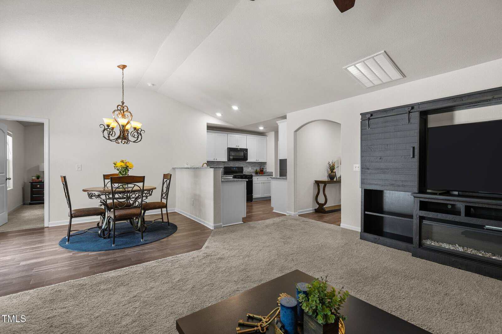 Open-concept living space featuring a carpeted living area with an entertainment unit, a dining area with a round table and chairs on a blue rug, and a kitchen with white cabinets and stainless steel appliances.