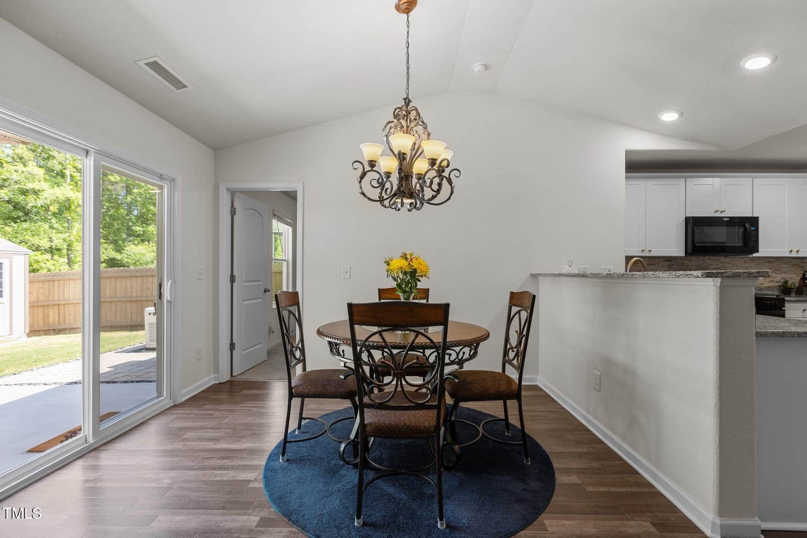 A dining area with a round table and four chairs on a blue rug, a vase of yellow flowers, a decorative chandelier, sliding glass doors to a fenced yard, and a view into a white kitchen.