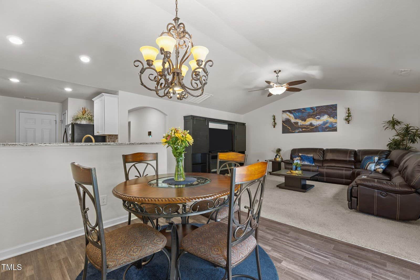A dining area with a round wooden table and four chairs sits next to a living room with a sectional sofa, coffee table, large TV, and wall art. A chandelier hangs above the table; kitchen is visible in the background.