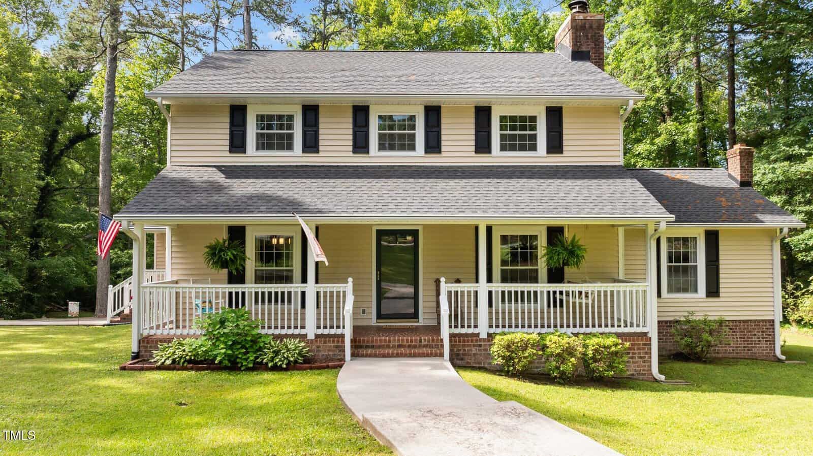 A two-story beige house with black shutters and a gray roof at 148 Shadowbrook Drive, Henderson, featuring a spacious front porch with white railings, hanging plants, and an American flag. The home is surrounded by green lawn and trees.