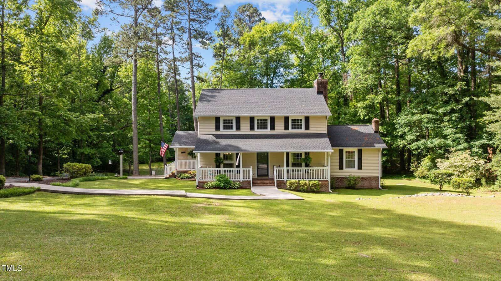 Two-story house with light-colored siding, dark shutters, and a covered front porch sits on a large, grassy lawn with a curved driveway, surrounded by tall trees under a bright sky.