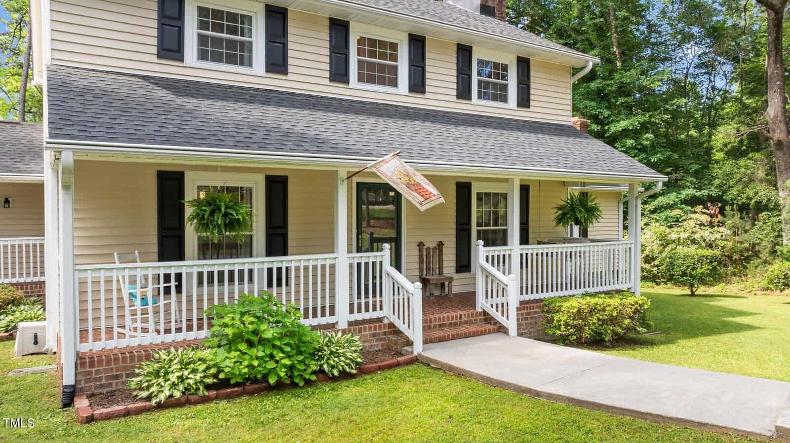 A beige two-story house with black shutters features a wide front porch with white railings, hanging ferns, a rocking chair, and an American flag. The porch overlooks a green lawn and is bordered by shrubs and trees.