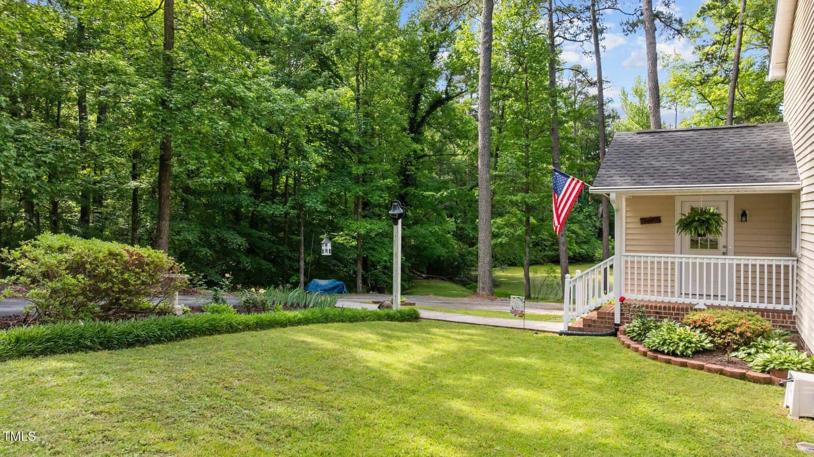 A neatly mowed front yard with a small porch, an American flag, and a hanging plant. The house is bordered by a wooded area with tall trees and a paved driveway leading to the street.
