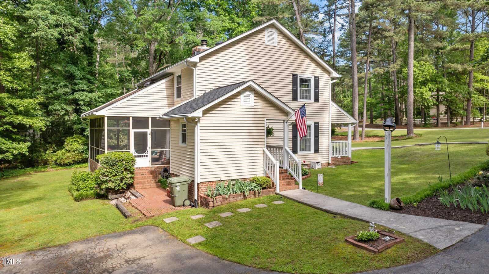 A two-story beige house with white trim, a screened porch, and an American flag on the front entry. The home is surrounded by grass, shrubs, trees, and a paved walkway leading to the entrance.