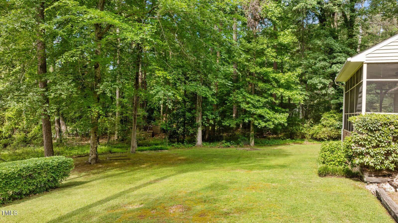 A grassy backyard bordered by a wooded area with tall green trees, next to a screened-in porch attached to a house on the right side of the image. Some low shrubs line the porch.