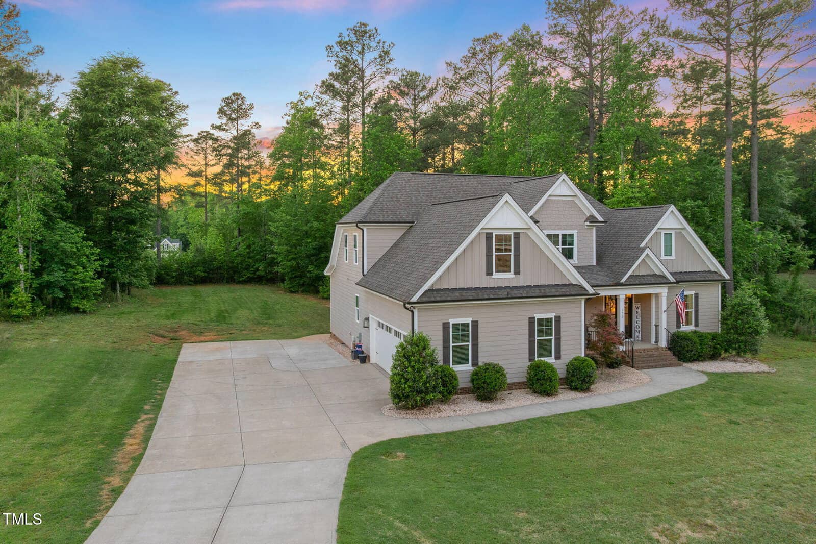Two-story beige house with a gray roof, multiple gables, and attached garage. Concrete driveway leads to the front entrance. The yard is grassy with bushes near the house, and trees surround the property at sunset.